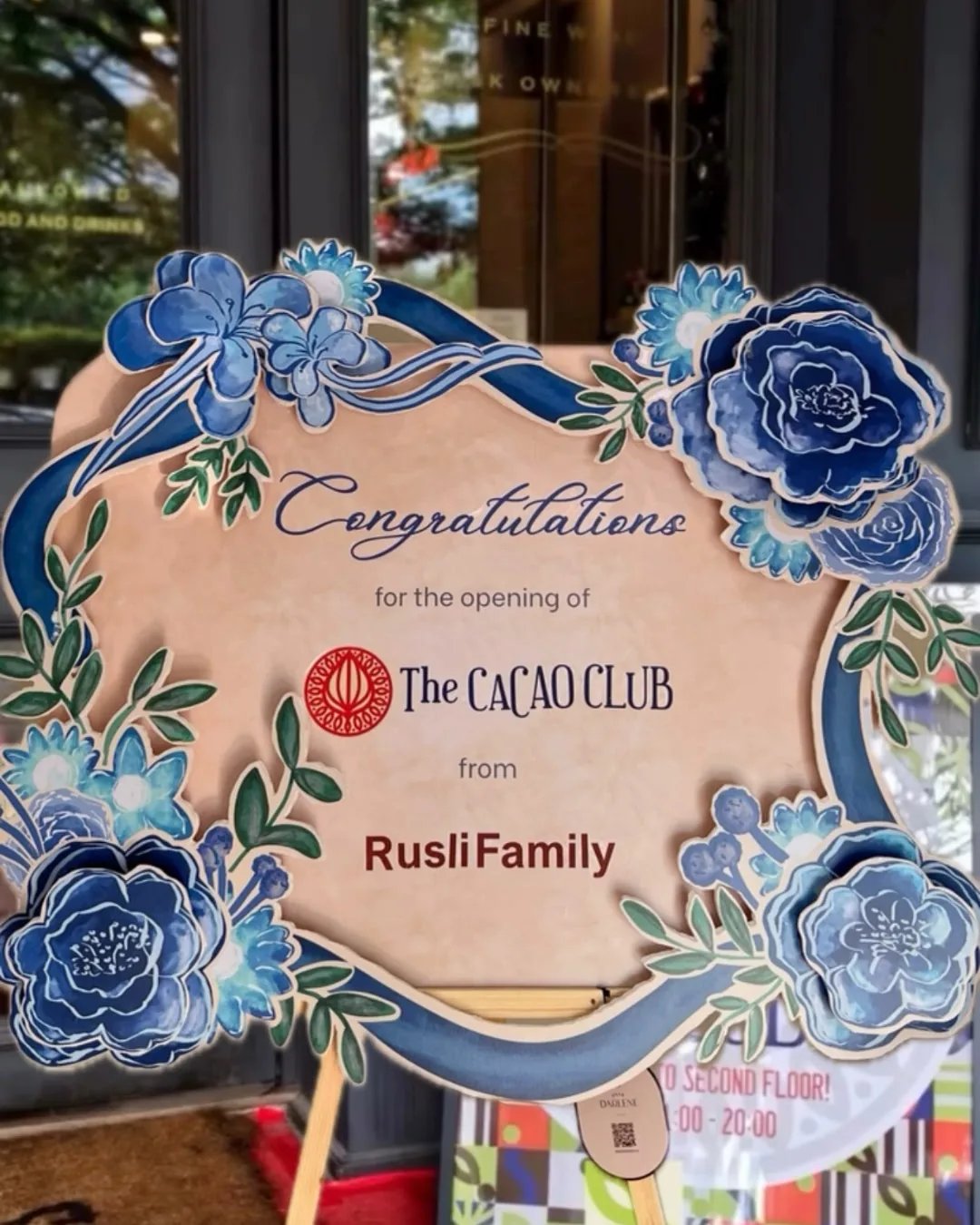 Papan ucapan The Cacao Club dengan ornamen bunga biru dan bidang tulisan berwarna krem.