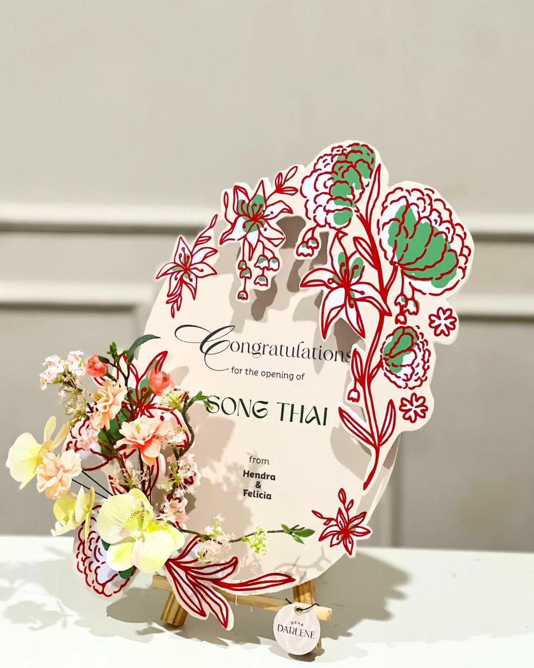 Papan ucapan Song Thai dengan ornamen merah hijau dan rangkaian bunga di sisi kiri.